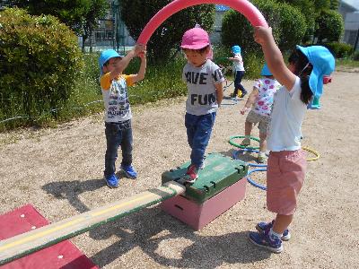 5歳児がアーチを手に持ち、その下の平均台を4歳児が渡っています。